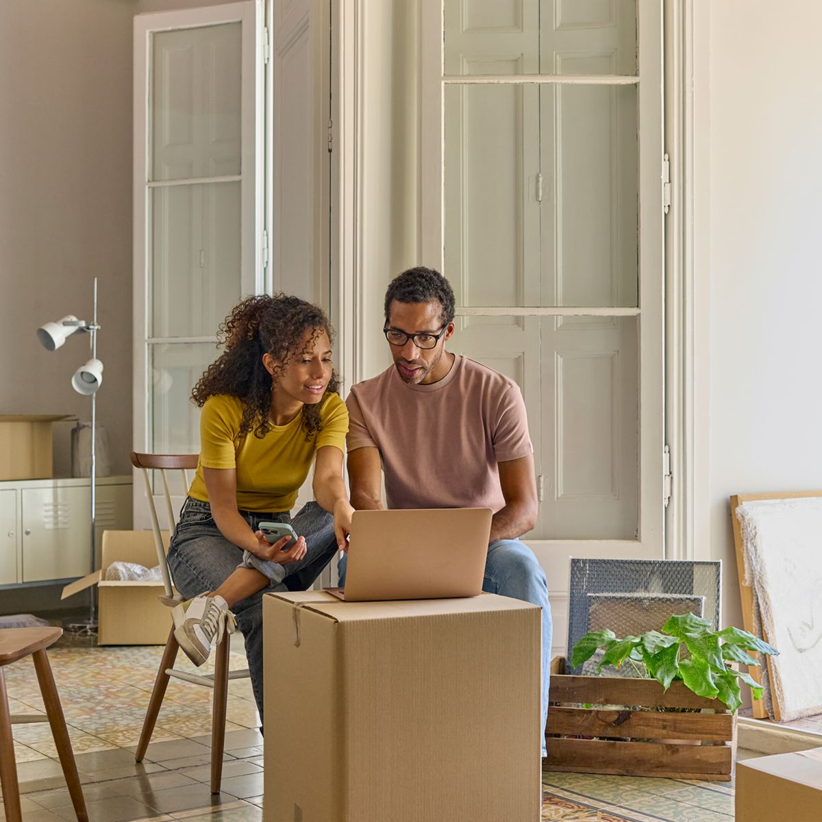 Two people sitting with a laptop and phone among moving boxes in a new home