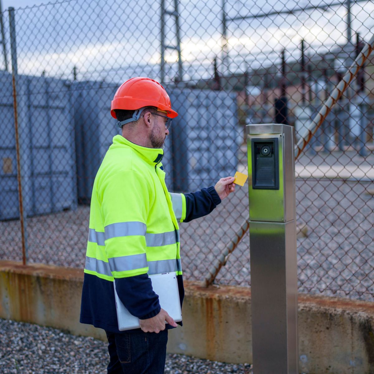 High-visibility jacket–clad utility worker holding a laptop swipes a key card at an access control reader beside a chain-link fence at an industrial site.