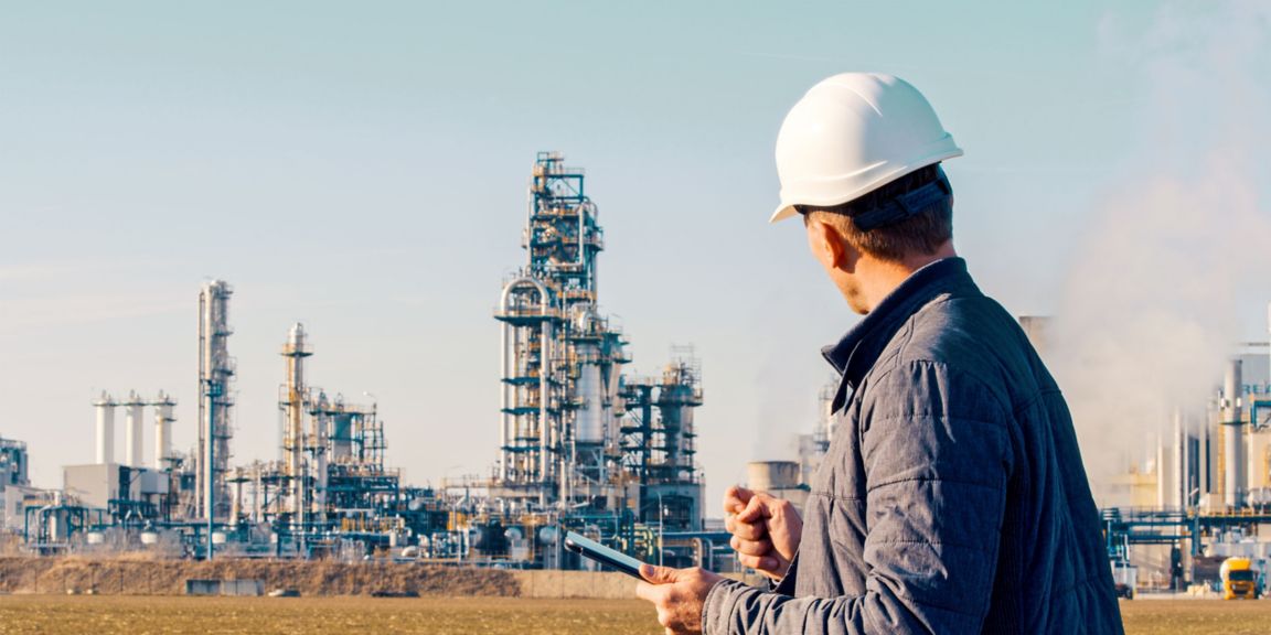 Worker wearing a hard hat uses a tablet while observing industrial processing equipment and infrastructure at a manufacturing facility.