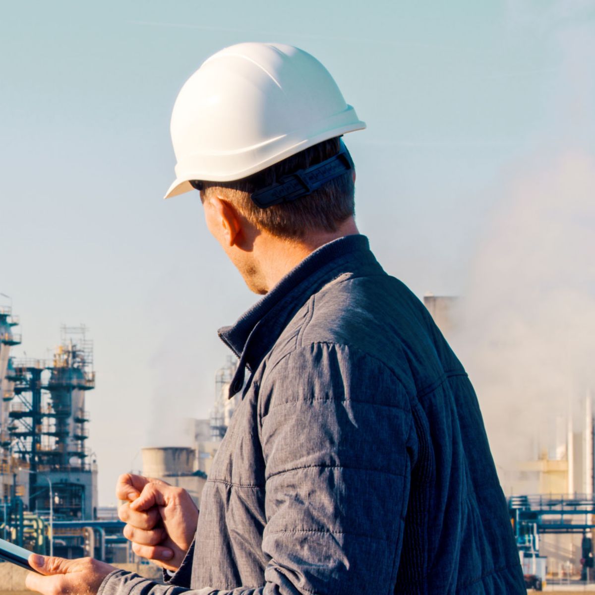 Worker wearing a hard hat uses a tablet while observing industrial processing equipment and infrastructure at a manufacturing facility.