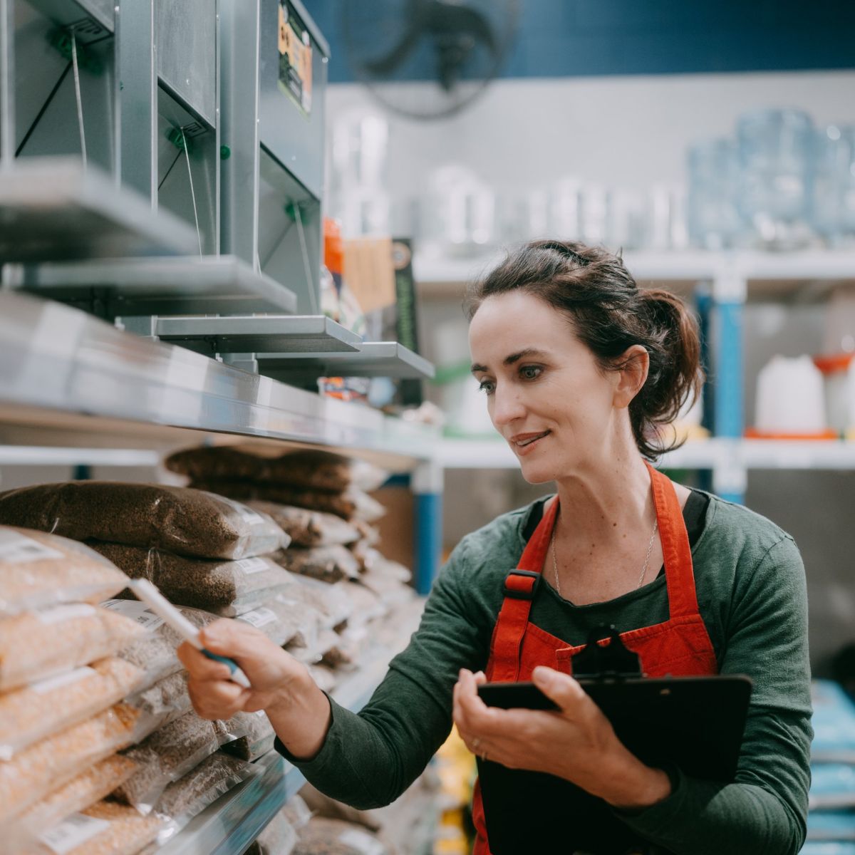 A store associate in a red apron conducts an inventory check of bagged goods in a retail warehouse setting.