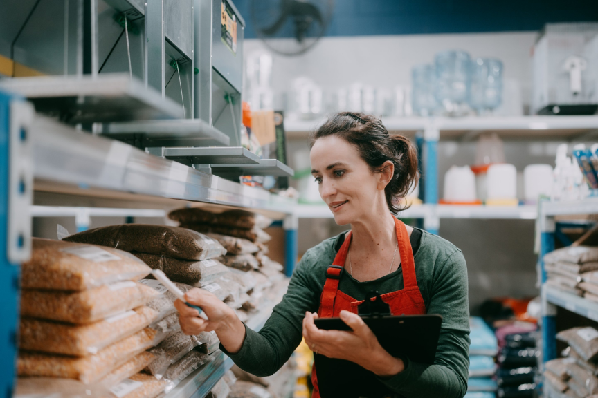 A store associate in a red apron conducts an inventory check of bagged goods in a retail warehouse setting.