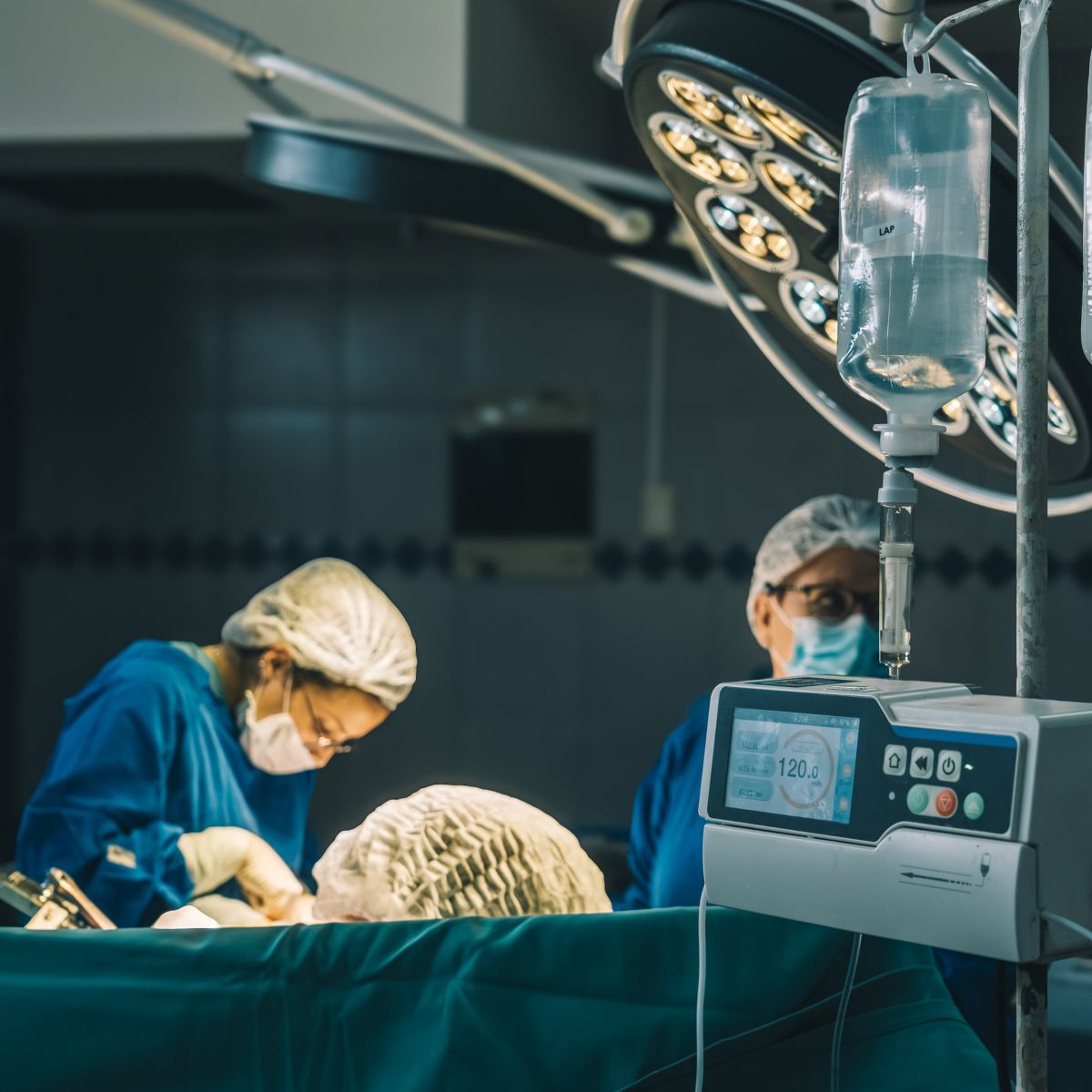 An infusion pump monitors intravenous fluid delivery while surgical staff operate in the background of a hospital theater.