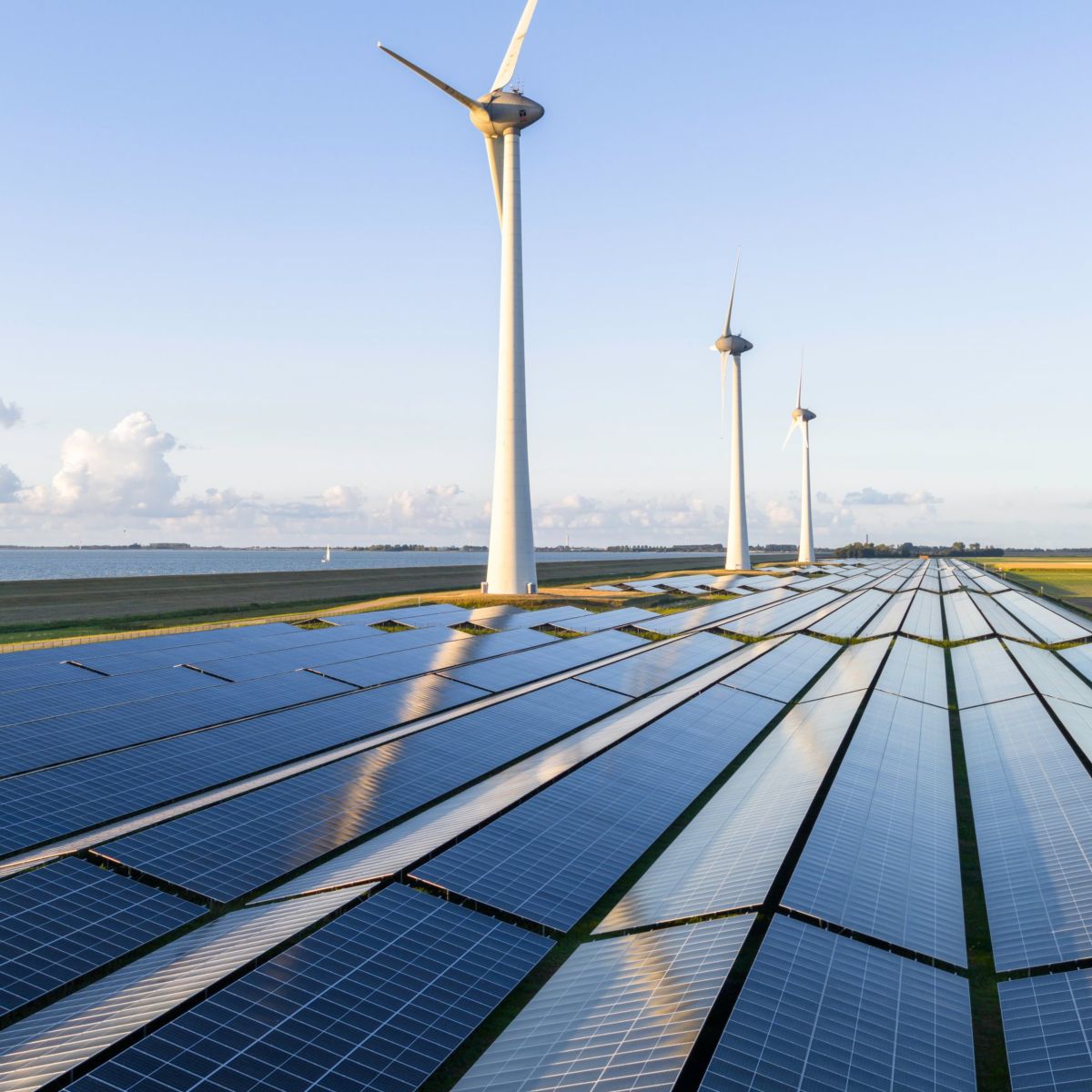 Aerial view of a solar farm integrated with wind turbines along a calm coastal landscape at sunrise.