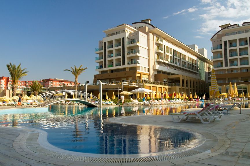 Luxury hotel resort exterior with a large swimming pool, sun loungers, umbrellas, and modern guest accommodation under a bright blue sky.