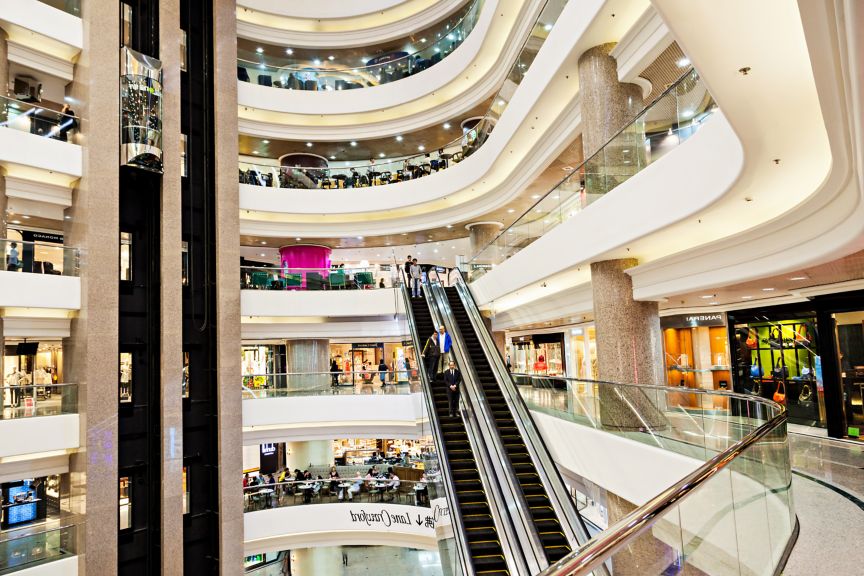 Multi-level shopping mall interior with escalators, retail stores, glass railings, bright lighting, and open commercial space.