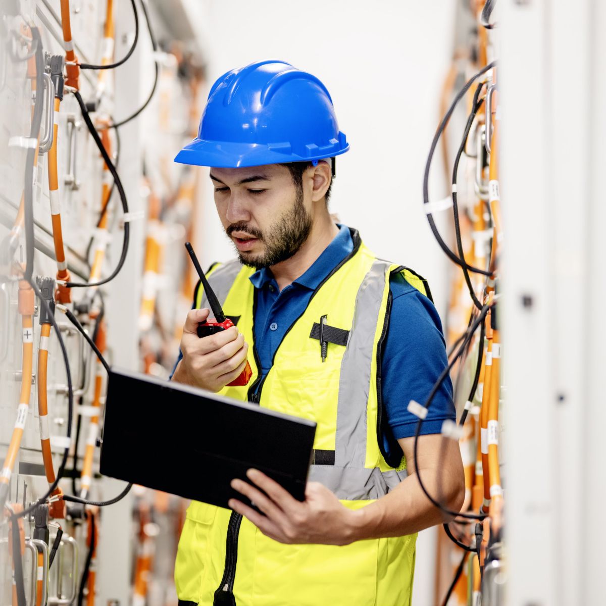 A technician in a safety vest and hard hat uses a tablet and two-way radio to monitor equipment in a server room facility.