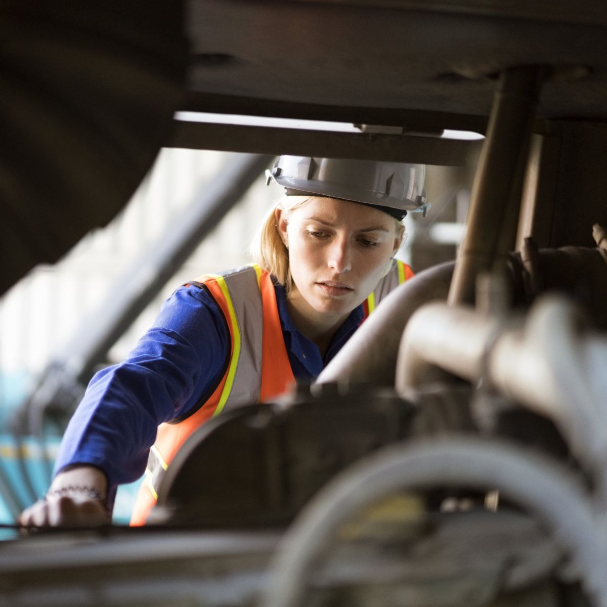 A female technician in a hard hat and safety vest performs a focused inspection of industrial machinery in a warehouse environment.