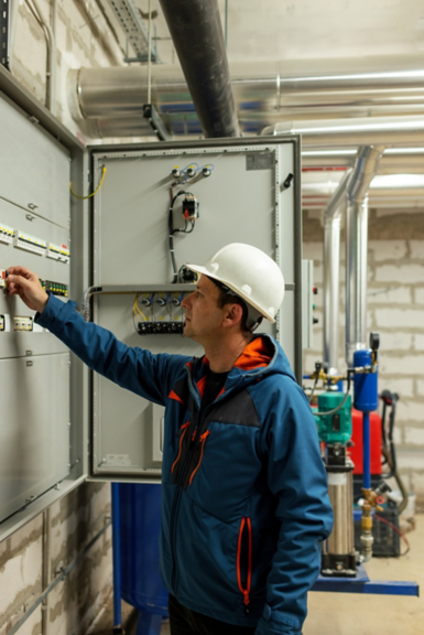 A professional technician in a hard hat inspects and operates an industrial electrical control panel inside a mechanical utility room.