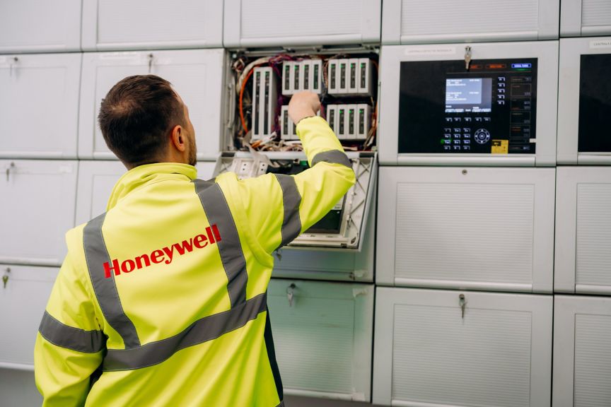 A Honeywell technician wearing a high-visibility jacket performs maintenance on an industrial electronic control panel in a technical server room.