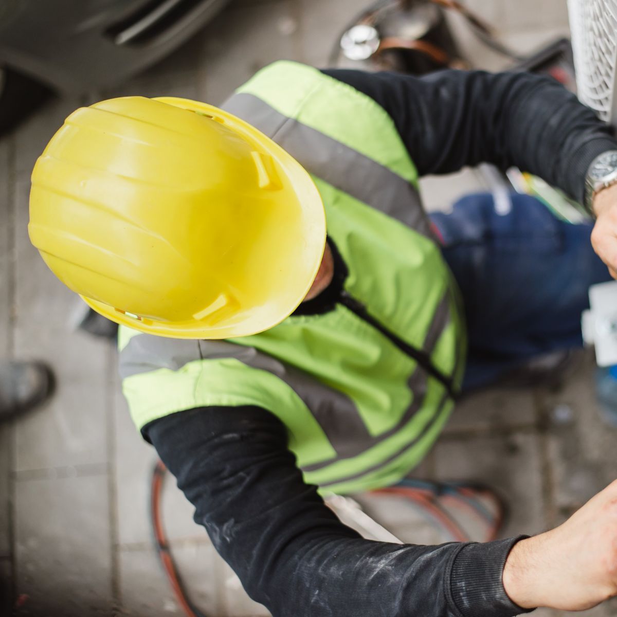 A professional technician in safety gear uses a wrench to repair an outdoor air conditioning unit's complex wiring and copper components.