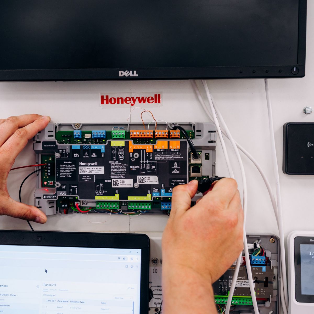 A technician installs wires into a Honeywell security control panel while configuring settings on a connected laptop.