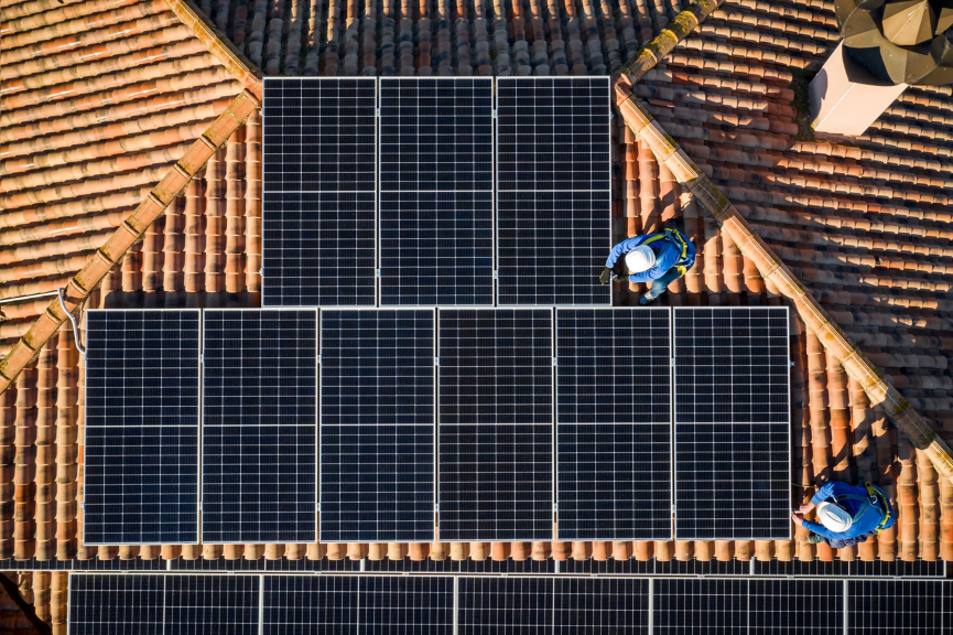 Aerial view of solar technicians installing photovoltaic panels on a traditional terracotta tile roof during a sunny day.