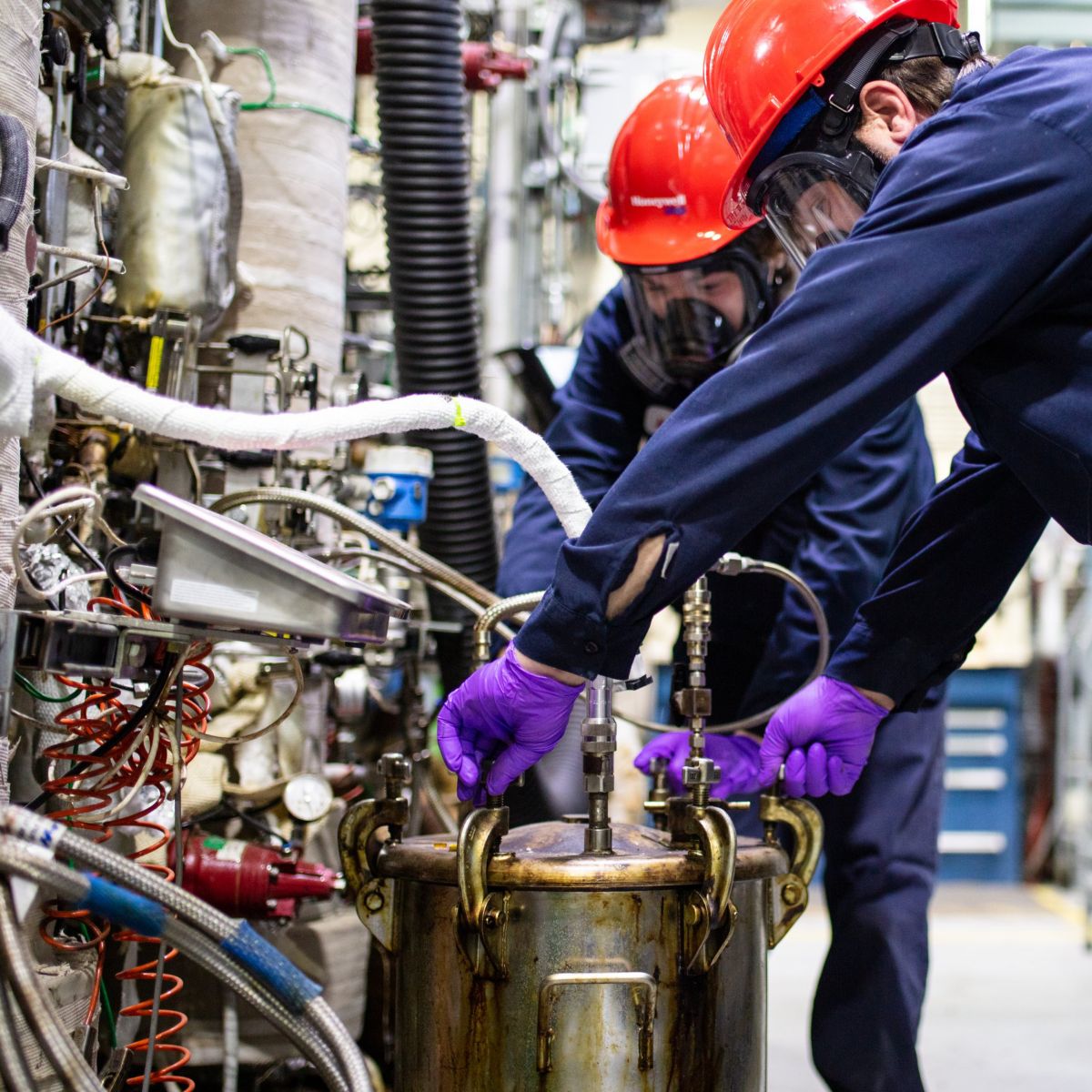 Two technicians in safety gear and purple gloves carefully adjust a pressurized metal vessel within a complex industrial manufacturing facility.