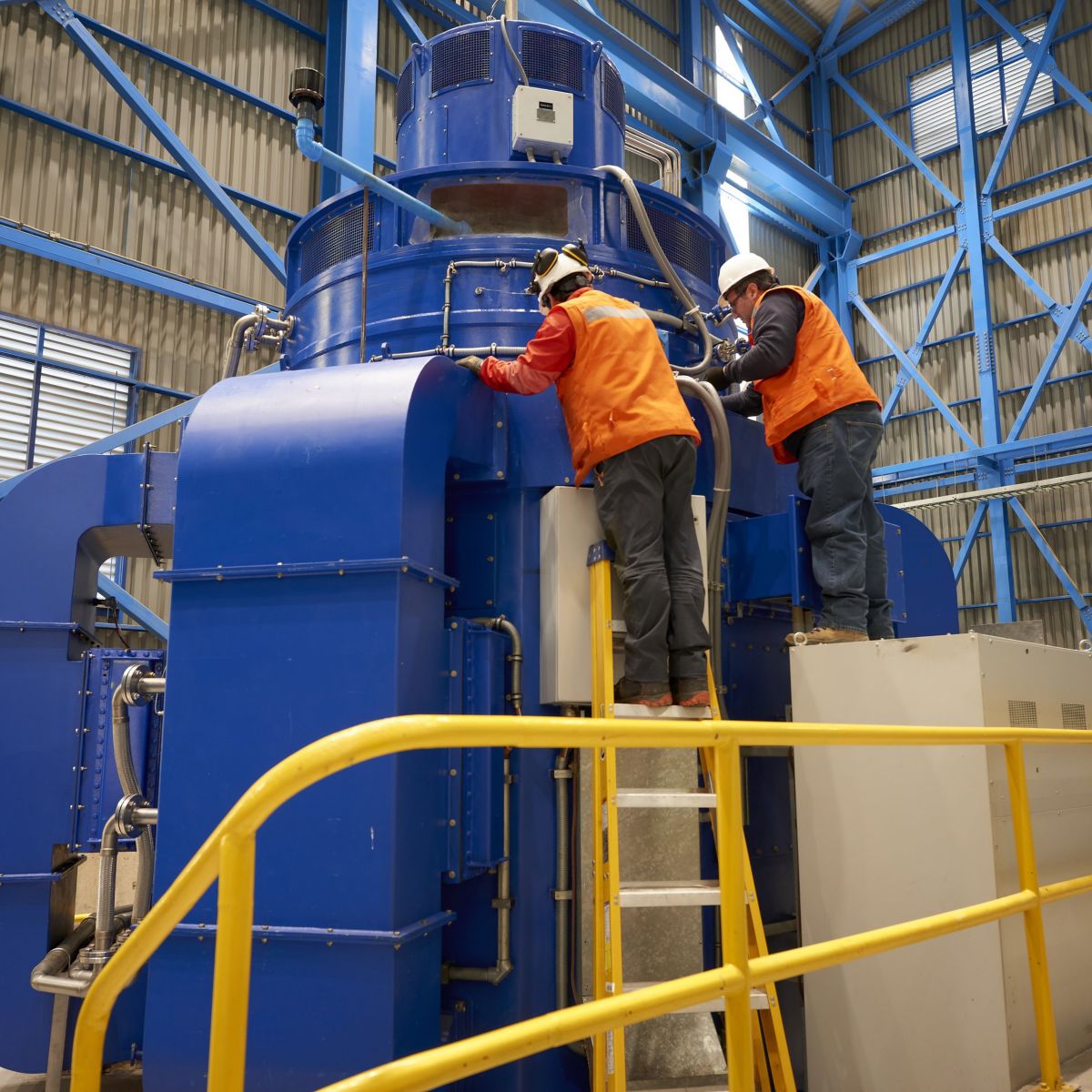 Two engineers in safety gear perform maintenance on a large blue industrial turbine within a spacious factory facility.