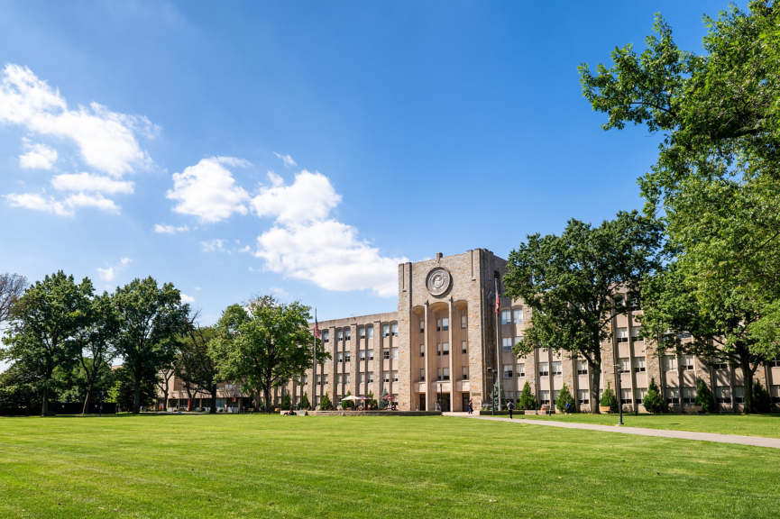 A stone collegiate building stands prominently behind a manicured green lawn under a bright blue sky.