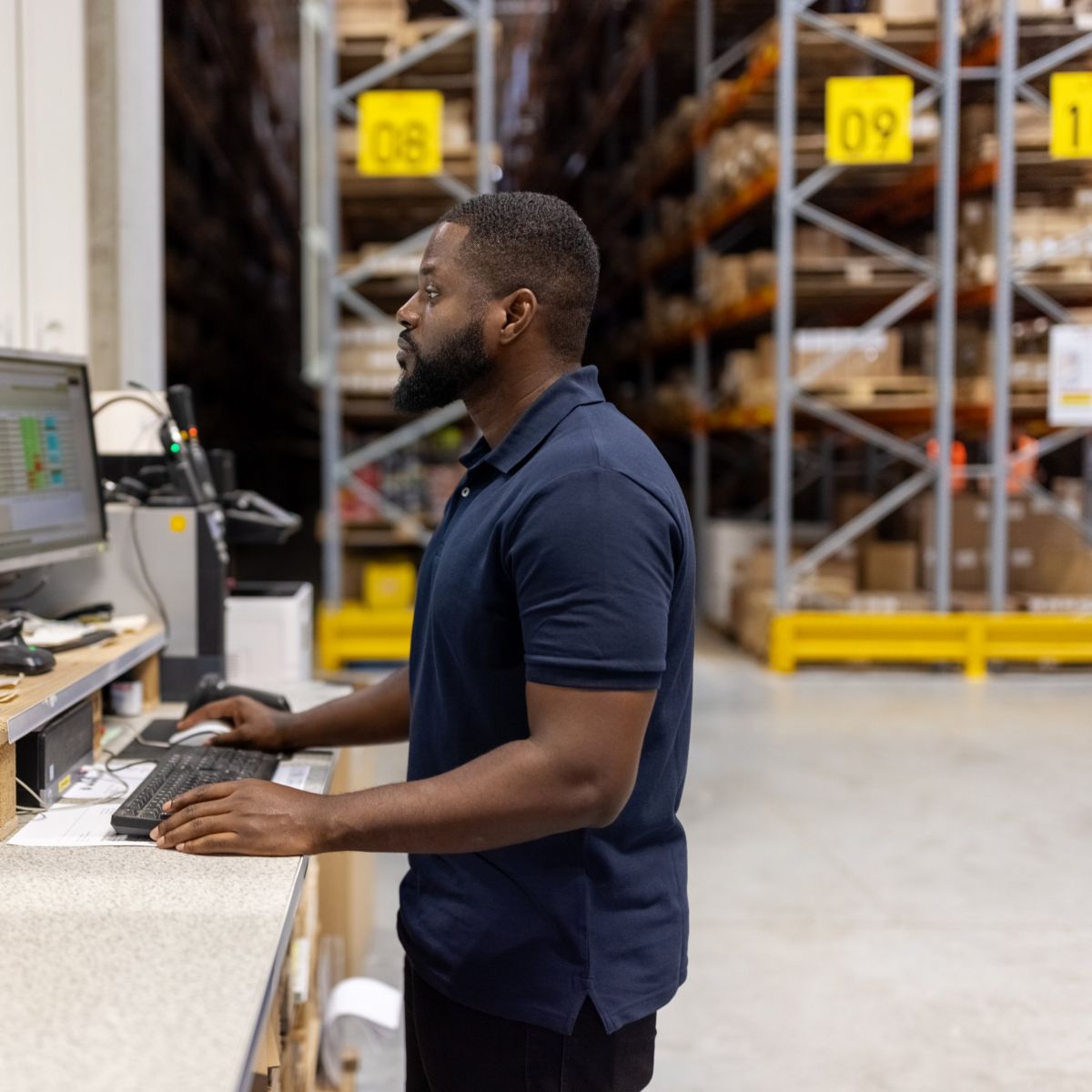 A warehouse worker monitors inventory and logistics data on computer screens at a packing station in a large distribution center.