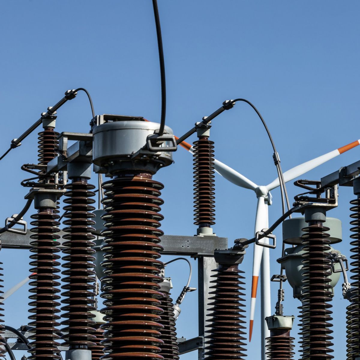 Close-up of industrial electrical insulators in a substation with wind turbines standing in the background against a clear blue sky.