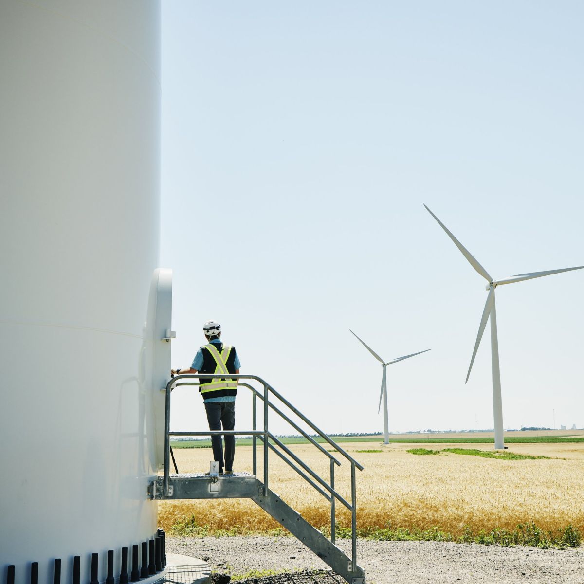 A technician in safety gear stands on a metal platform to access a large white wind turbine tower situated in a vast golden wheat field.