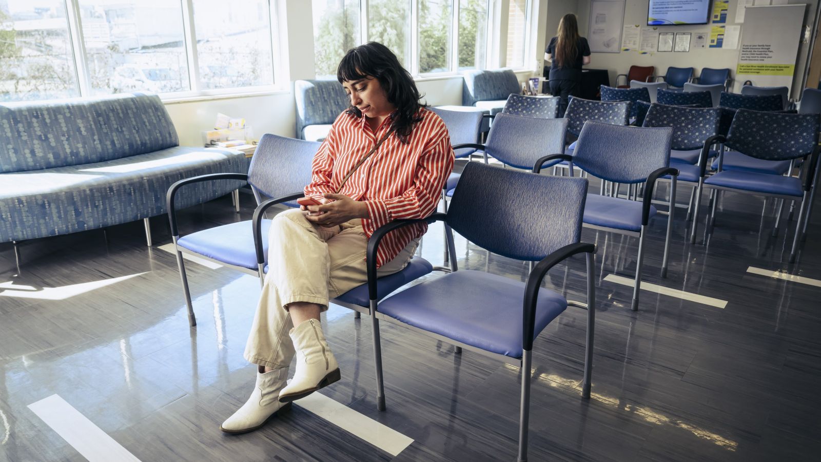 A woman in a striped shirt sits in a brightly lit office waiting area while looking at her smartphone.