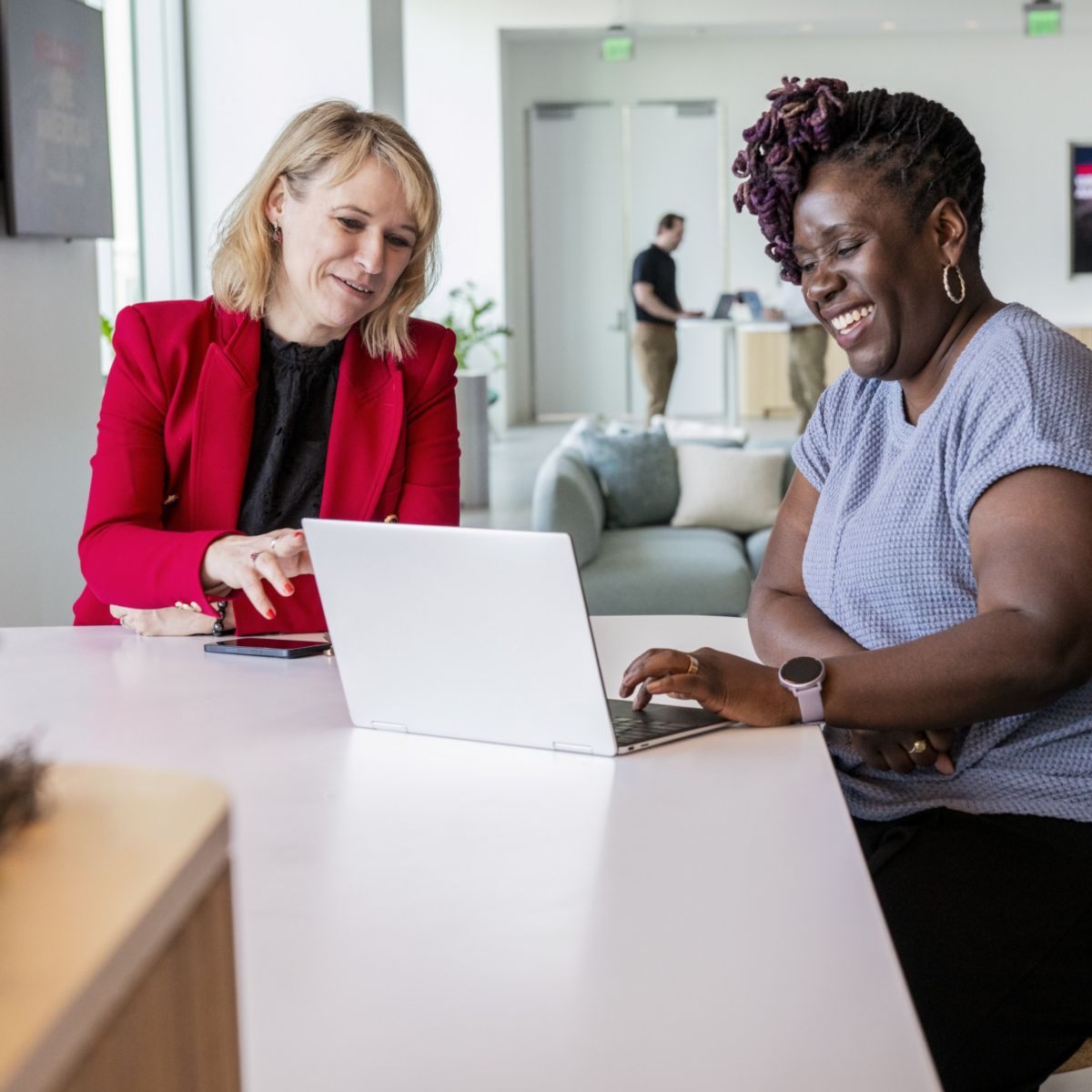 Two people sitting at a table using a laptop, with a phone nearby, in an indoor office space with seating in the background.