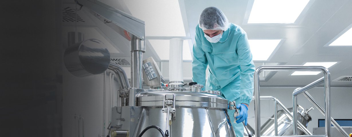 A masked worker in teal protective clothing operates a large stainless-steel pharmaceutical vessel.