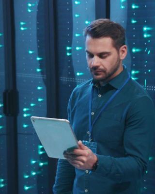 IT professional standing in a data center with illuminated server racks, holding a tablet and analyzing system information.