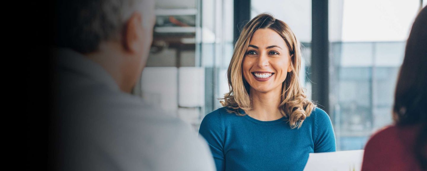 Smiling woman in a blue shirt holding papers while talking with two colleagues in an office meeting, symbolizing communication and collaboration.