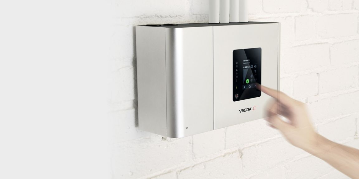 A hand is about to touch a control button of a VESDA panel mounted on a white wall.