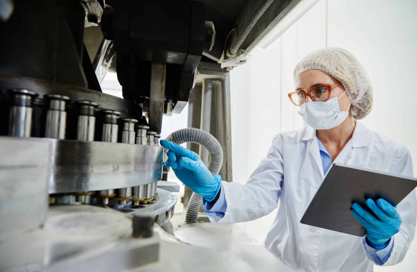 Professional female process technician in vinyl gloves and mask inspecting machinery while pointing at punch tools of tablet press machine at pharmaceutical manufacturing plant