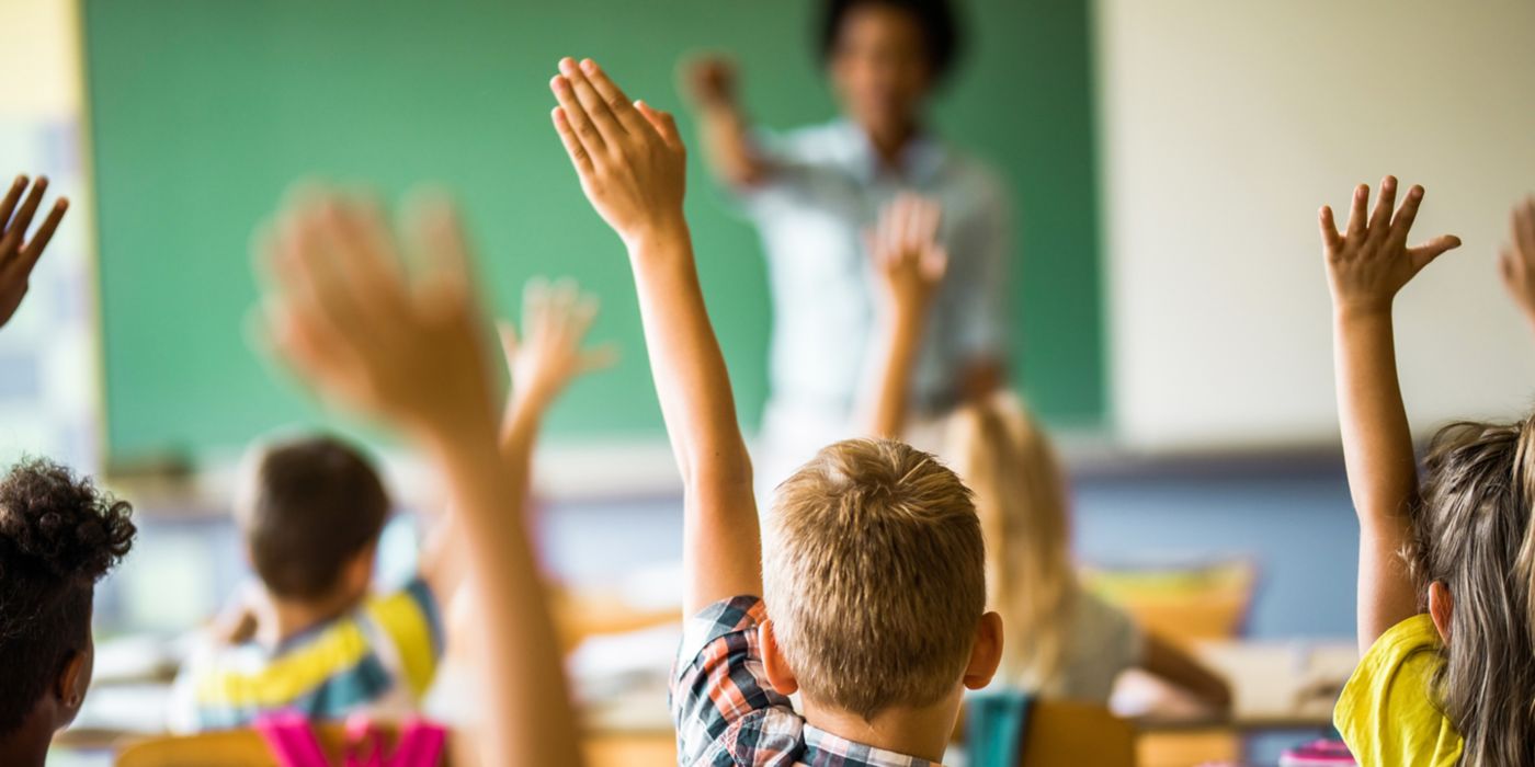 Rear view of large group of students raising their hands to answer the question on a class at elementary school.