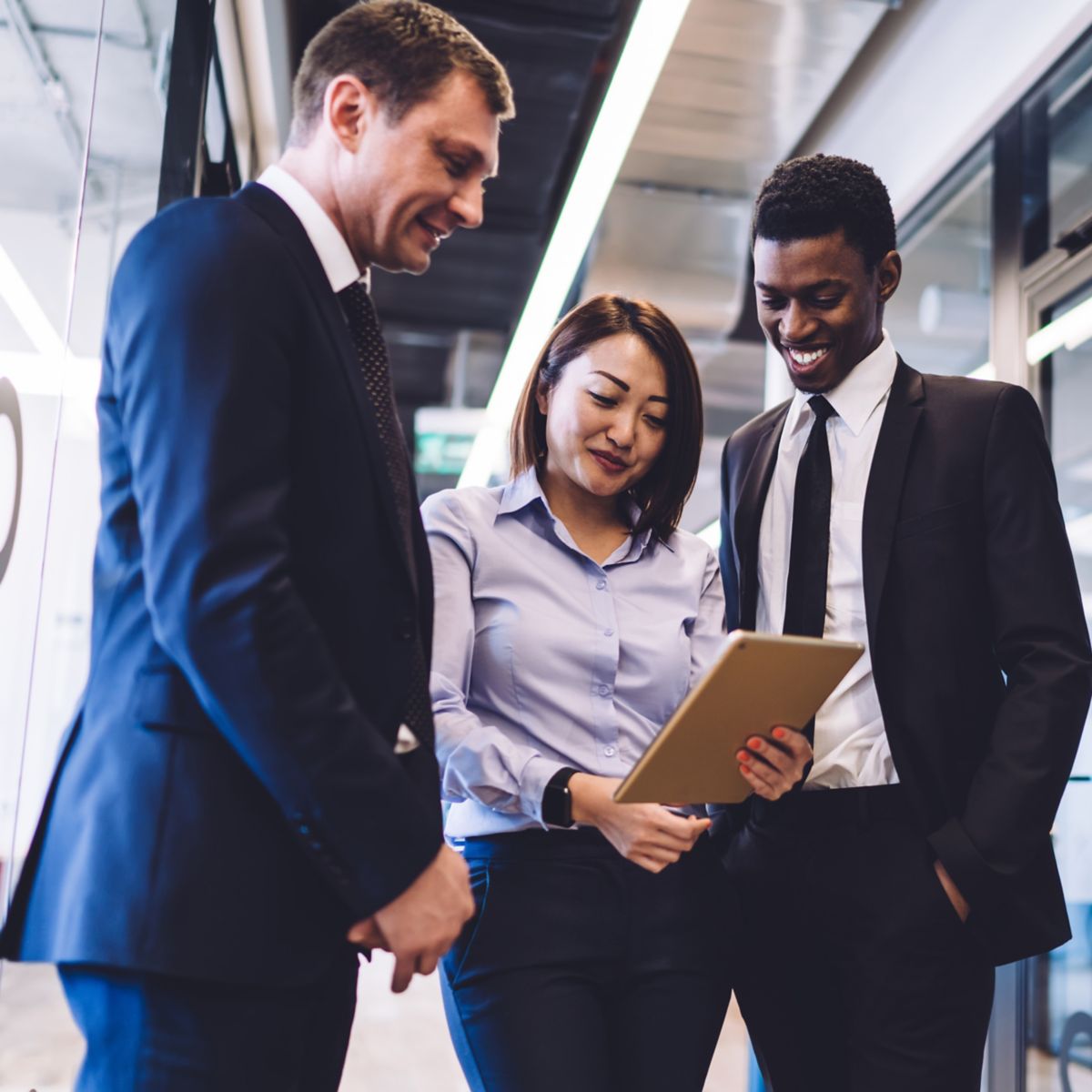 Asian young woman with wristwatch holding tablet and standing with African American male with toothy smile and cheerful businessman in formal suit