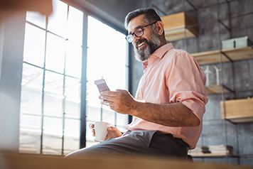 Mature businessman working at modern office space. Sitting at the table, using phone . Holding cup of coffee.