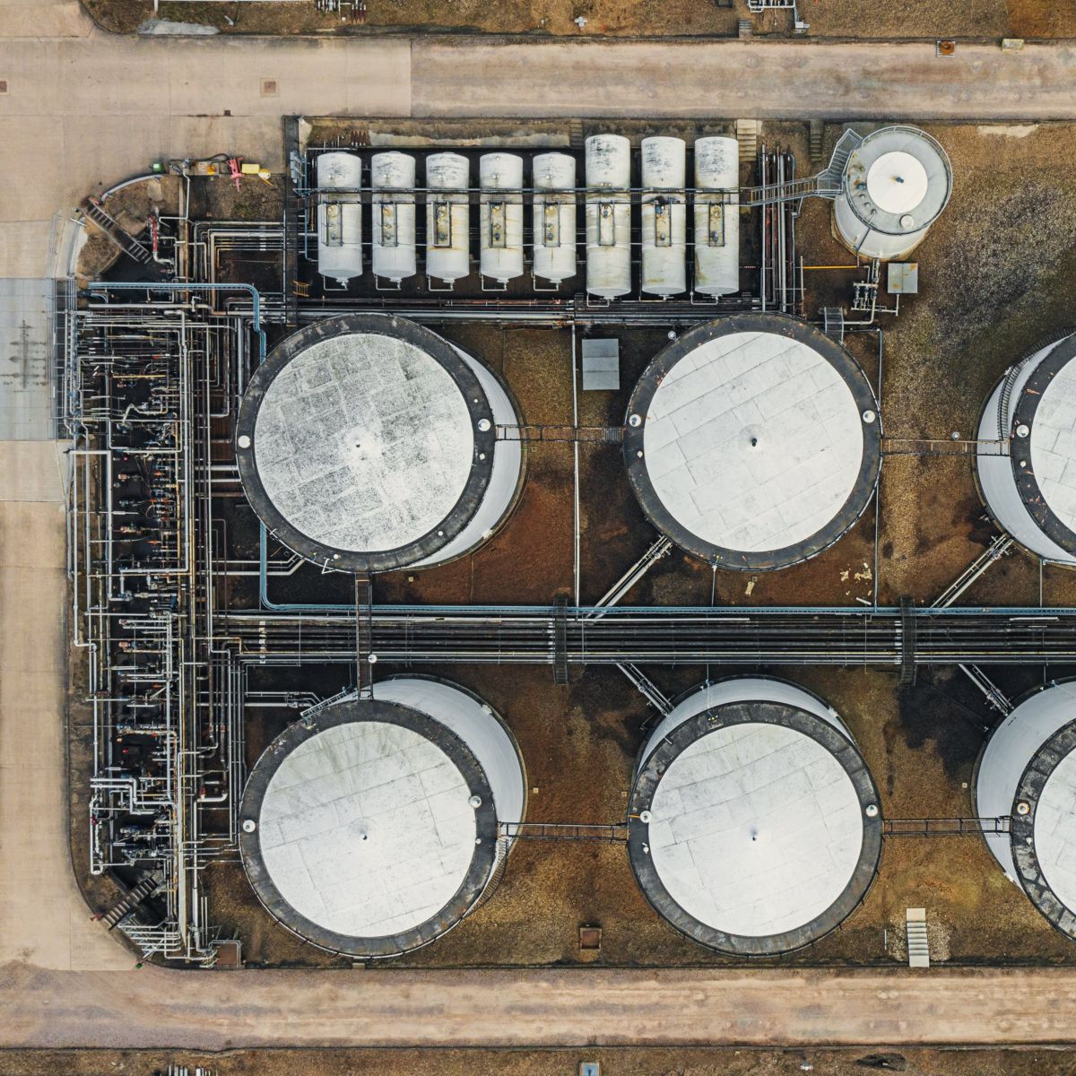 An overhead view of an industrial terminal featuring large white storage tanks and complex pipeline networks situated on a gravel site.