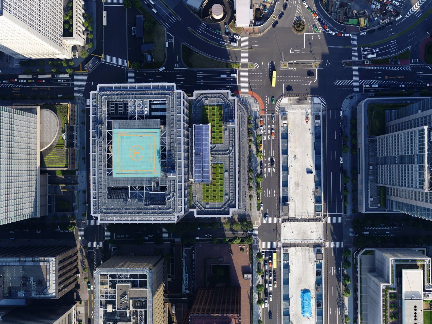 A high-angle aerial view of a dense Tokyo urban intersection featuring high-rise buildings, rooftop helipads, and organized road traffic flow.