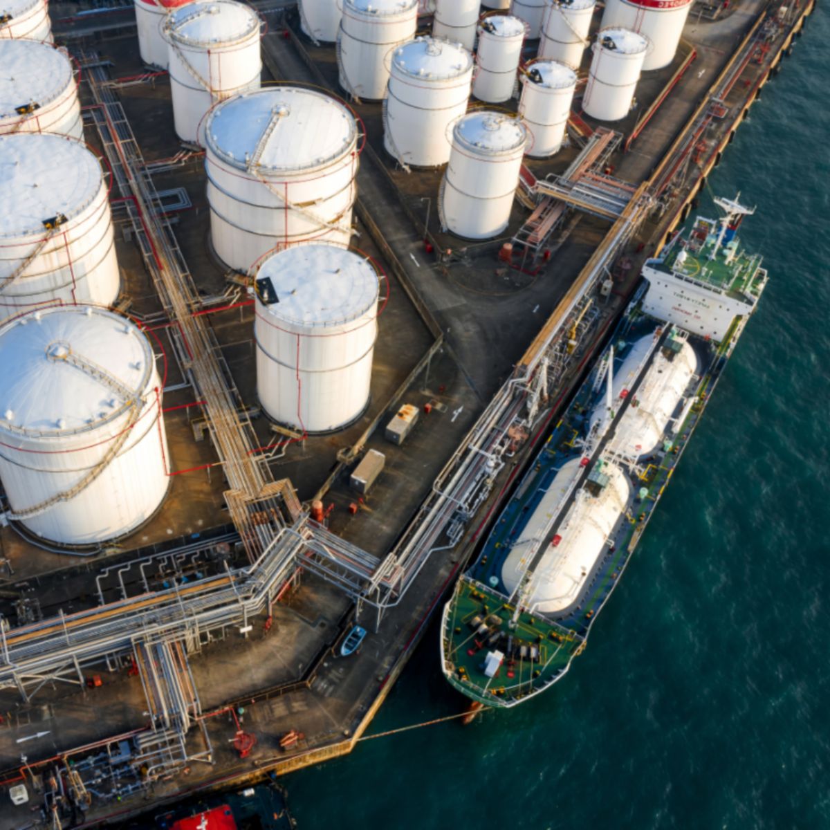 Industrial site with white storage tanks, red bands, and a docked ship equipped with cylindrical tanks.