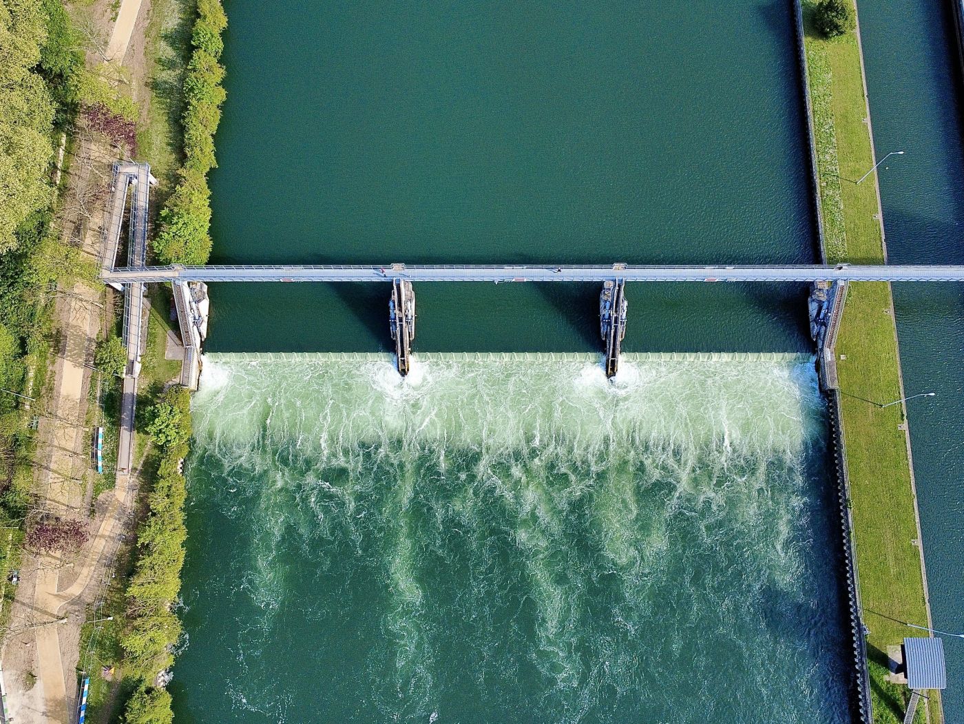 An overhead aerial view shows water flowing over a concrete weir structure on a canal, framed by green riverbanks and a pedestrian access bridge.