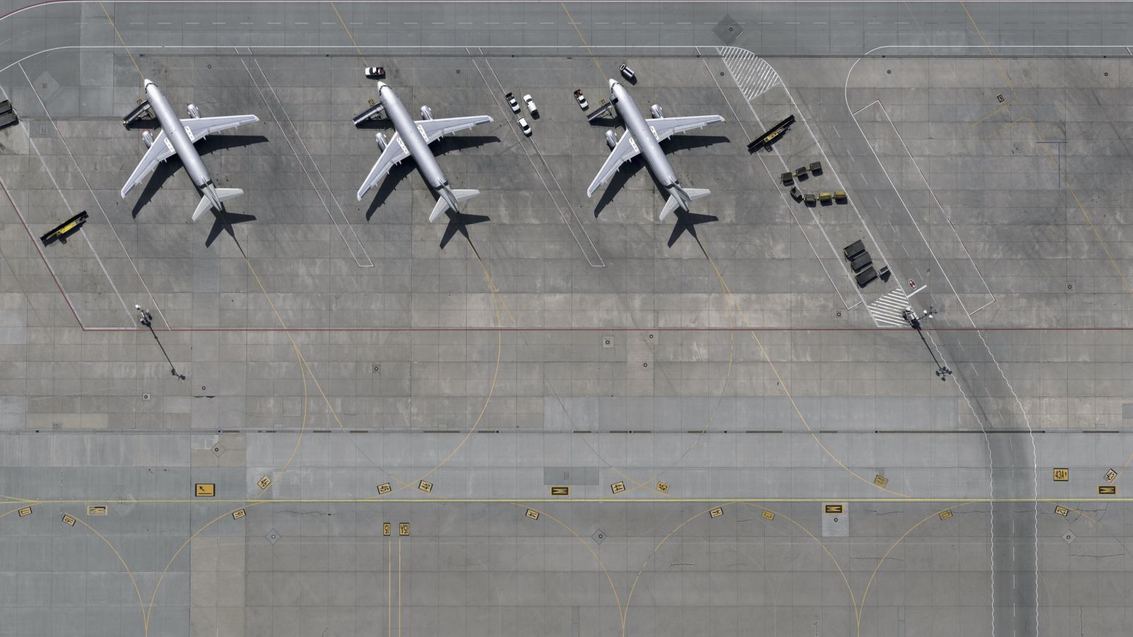 An aerial perspective shows three passenger planes parked at an airport terminal surrounded by ground support equipment and runway markings.