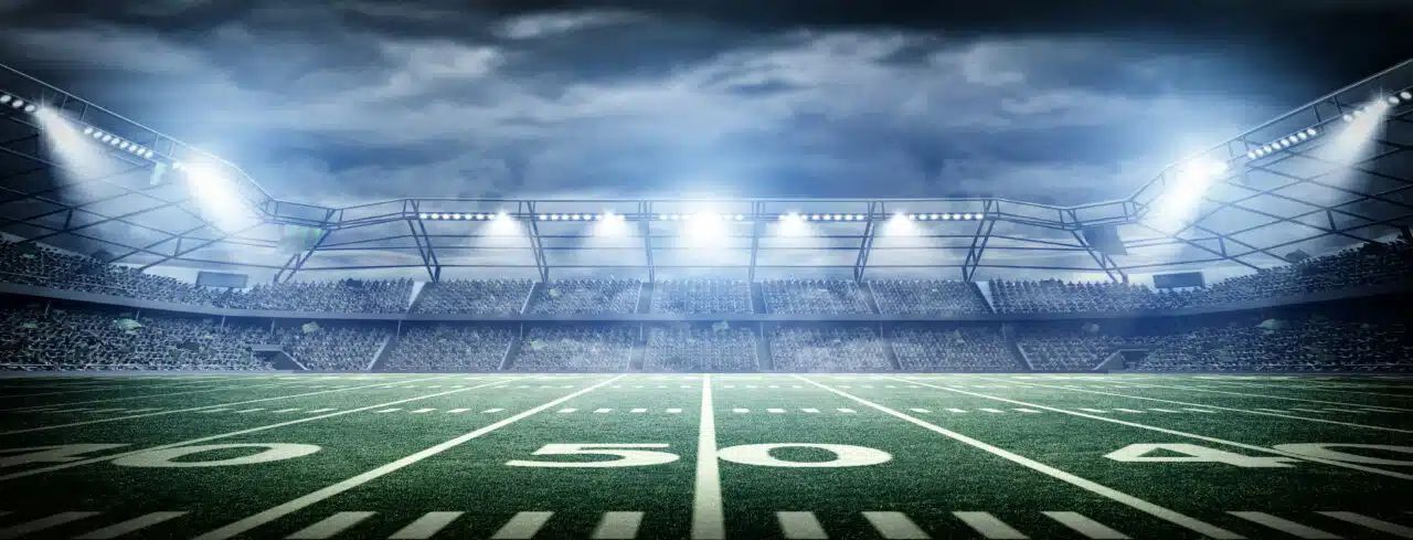 Illuminated American football stadium with field markings and spectator stands, representing a large-scale sports venue environment.