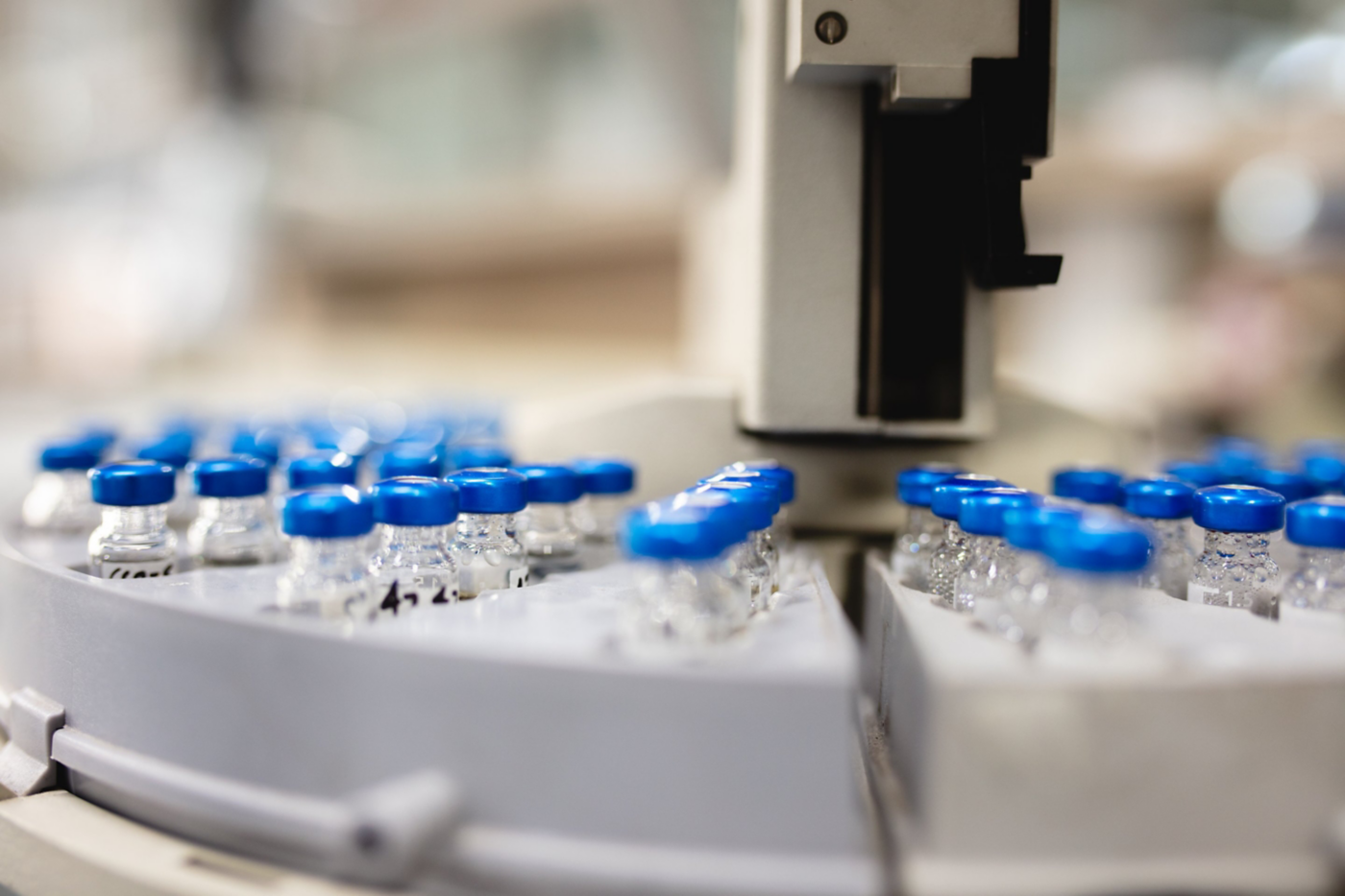 A robotic arm samples a carousel of sealed medical vials during an automated quality control inspection in a sterile laboratory environment.