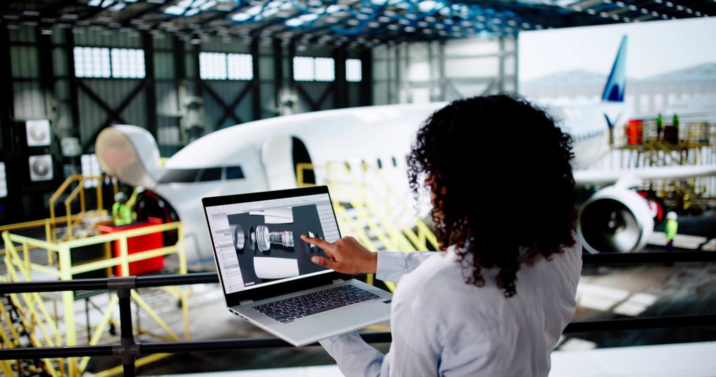 An engineer reviews aircraft component 3D models on a laptop while overlooking a commercial jet undergoing maintenance in a large hangar.