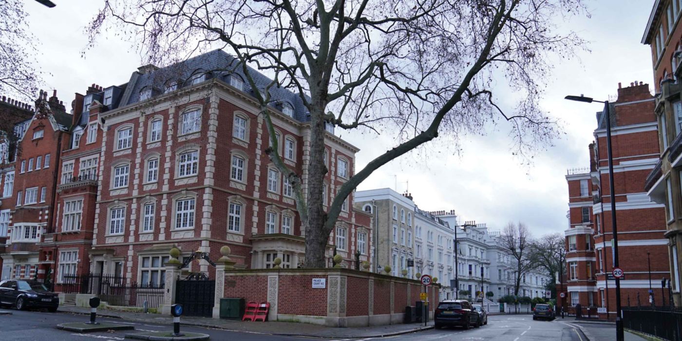  A quiet residential street corner featuring multi-story brick buildings with white-framed windows and ornate architectural details. A large tree and parked cars line the street under an overcast sky, creating a calm urban neighborhood scene.