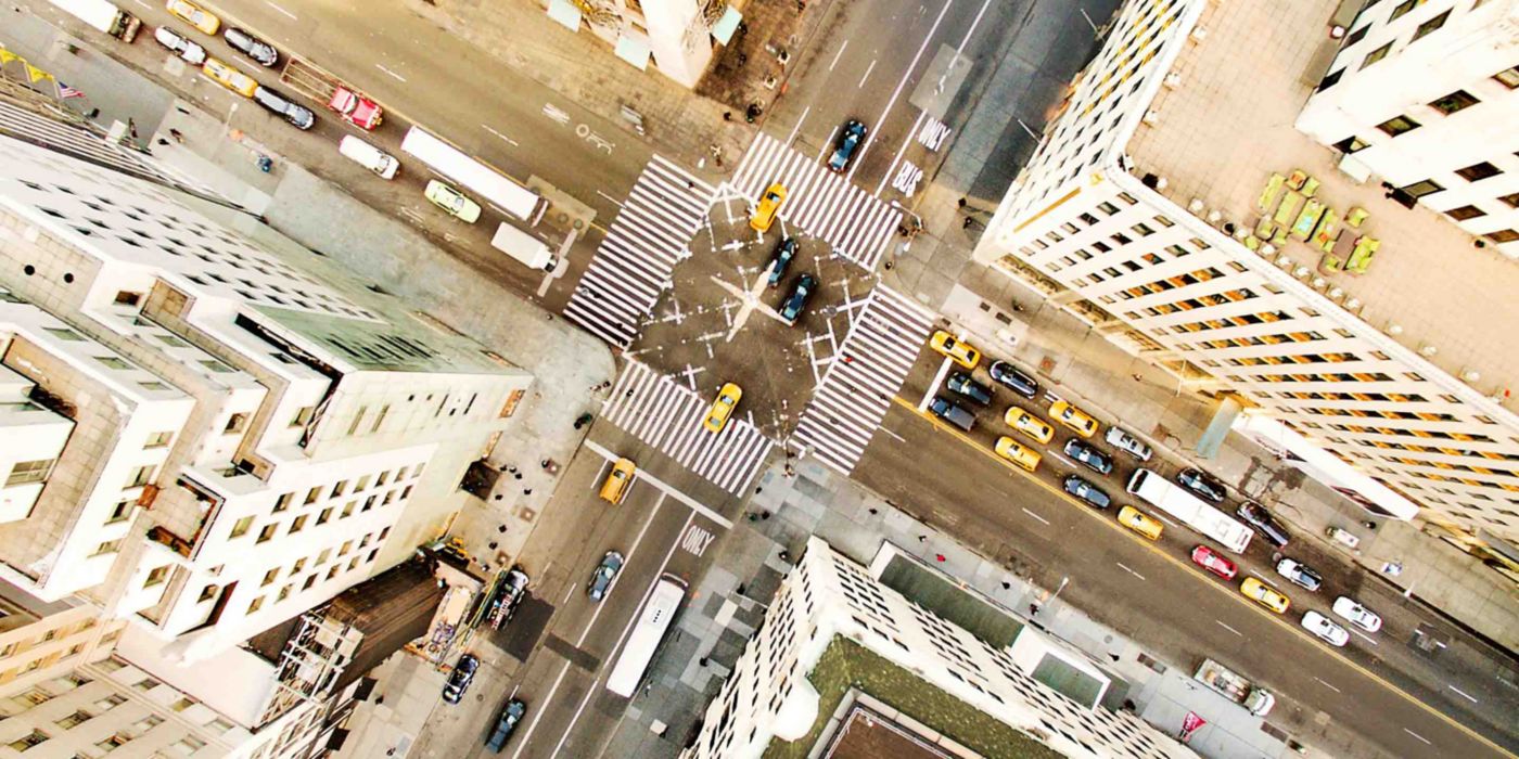 An overhead view of a bustling urban intersection featuring yellow taxis, cars, and buses navigating multiple lanes and crosswalks. Tall buildings surround the area, with rooftops and street markings clearly visible.