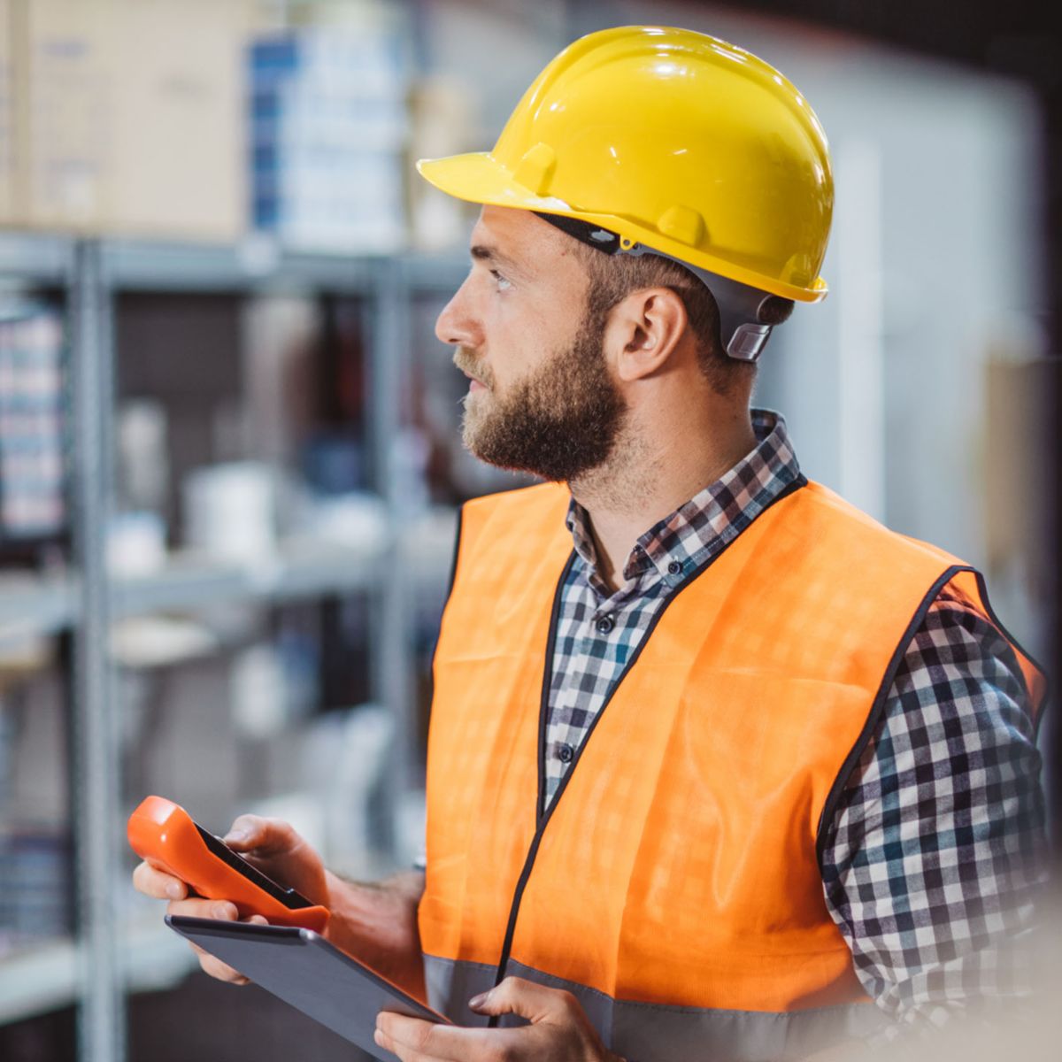 A warehouse employee wearing a yellow hard hat and orange safety vest holds a clipboard and handheld device while inspecting inventory. The background features shelves stocked with various items, highlighting a safe and organized work environment.