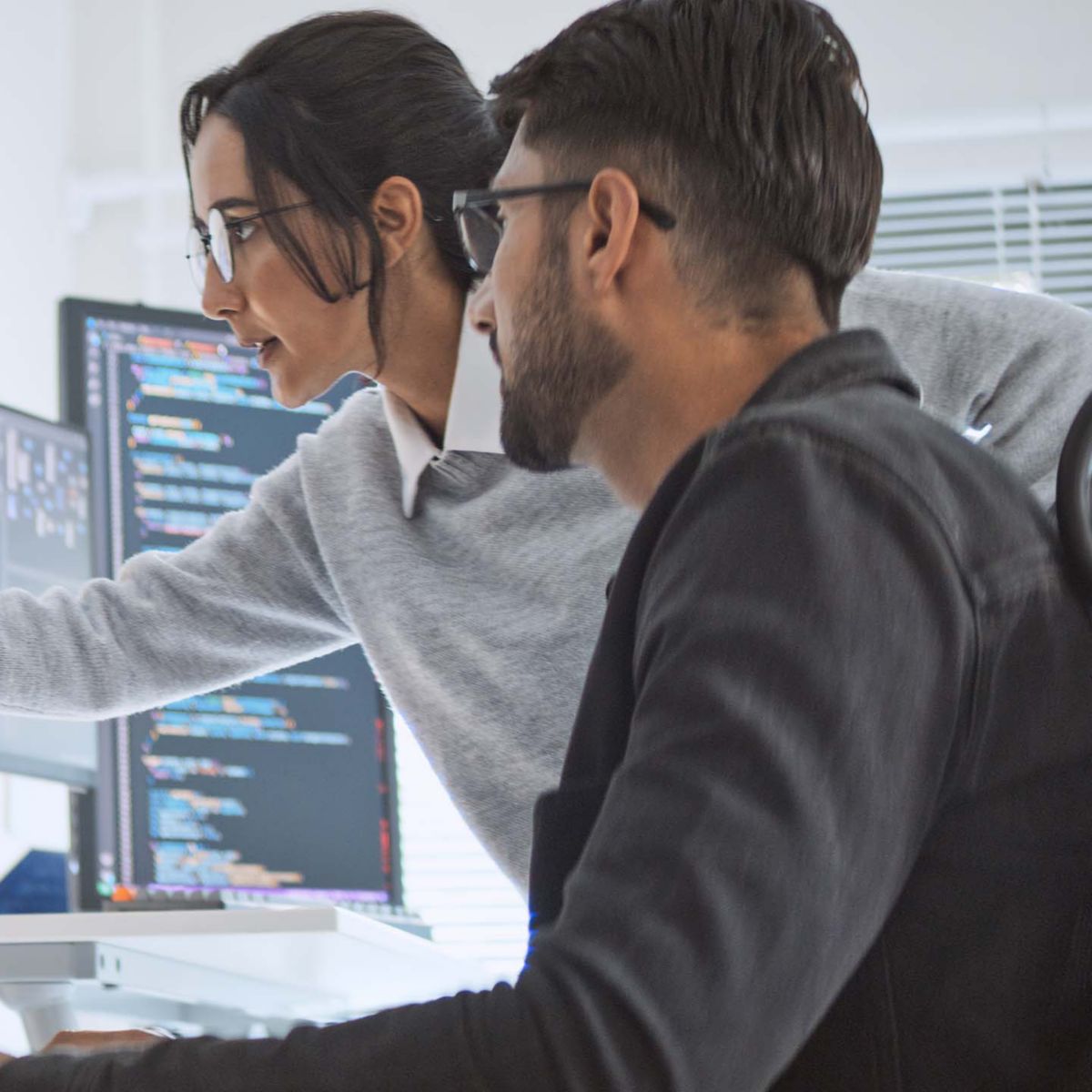 Two individuals work together at a desk with multiple monitors displaying code. One points at the screen while the other observes, highlighting teamwork in a bright, modern office environment with natural light filtering through blinds.