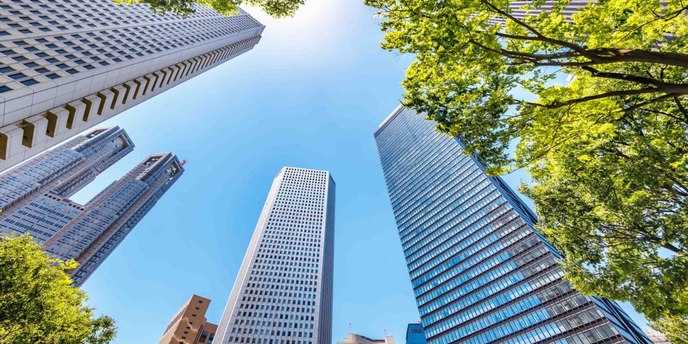 An upward view of tall skyscrapers surrounded by lush green trees, set against a bright blue sky with the sun shining overhead. The image captures the harmony between urban architecture and nature.