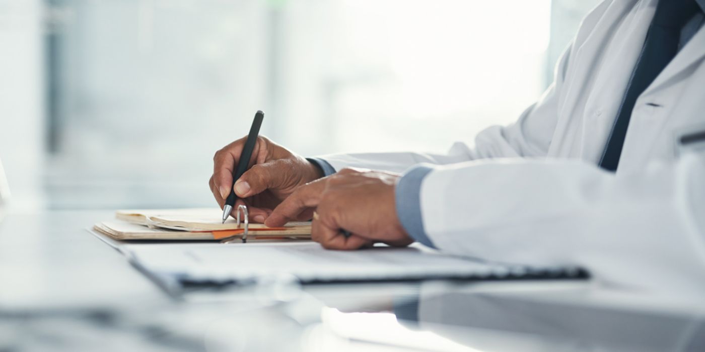 A close-up of a medical professional in a white coat writing on a clipboard or notepad. The desk includes documents that appear to be medical records, emphasizing clinical documentation and healthcare practice.