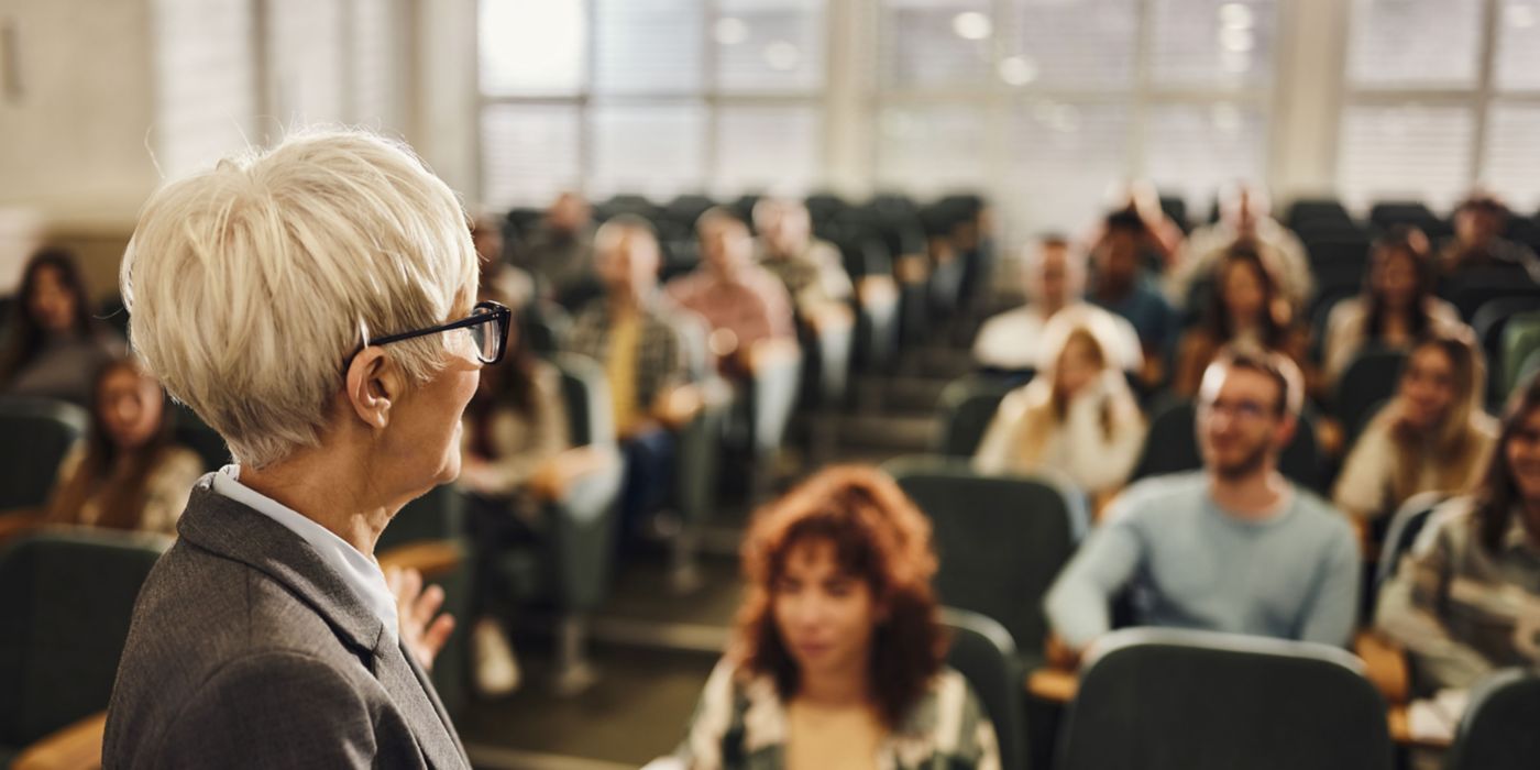 A classroom or lecture hall scene featuring a professor or lecturer standing at the front, speaking to an audience of students. The image captures the academic environment with natural light filtering through windows and students attentively facing forward.