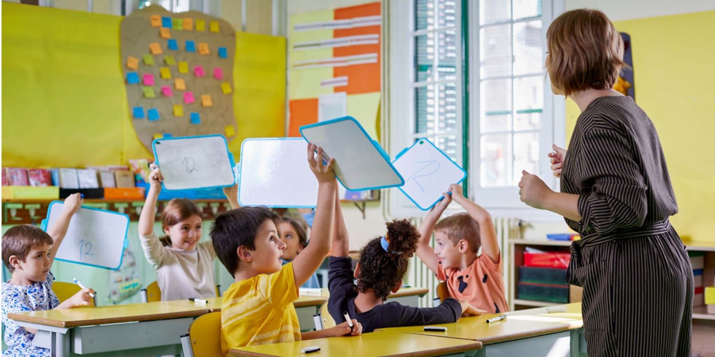 Children show numbers on whiteboards while teacher watches attentively.
