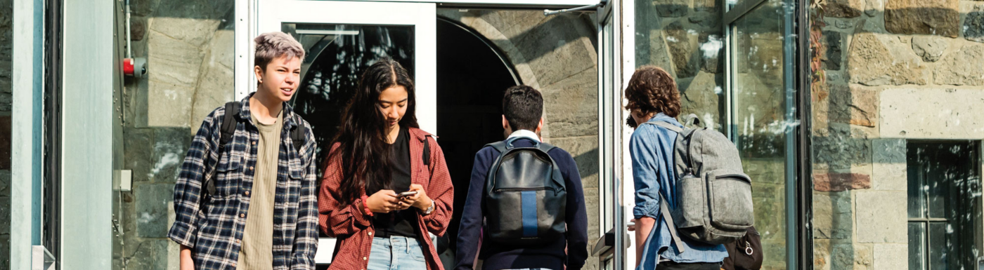 Four individuals, some with backpacks, stand near a modern building entrance with glass doors and a stone exterior. Ideal for illustrating campus life, office entry, or group activity in urban settings.